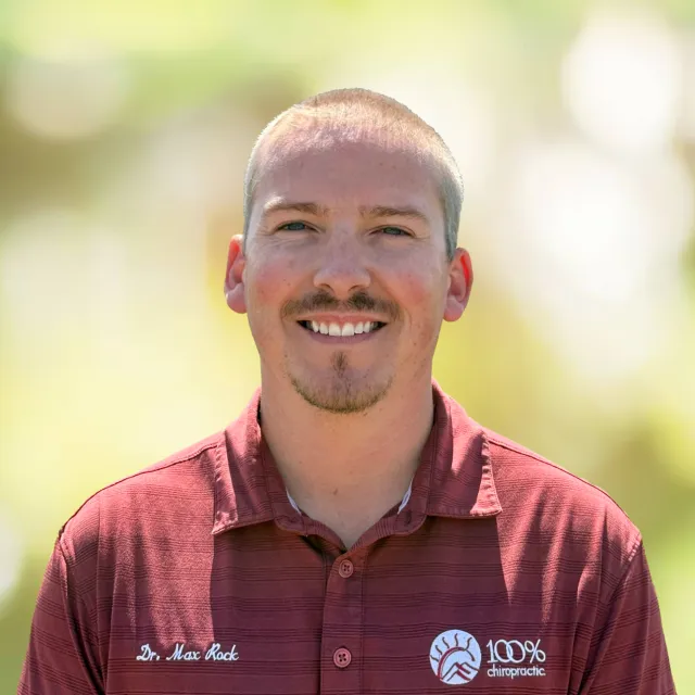 Smiling man in a maroon 100% chiropractic shirt with name Dr. Max Rock outdoors with blurred green background