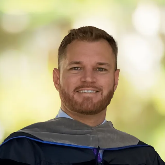 Smiling graduate with beard wearing academic hood and robe against blurred outdoor background