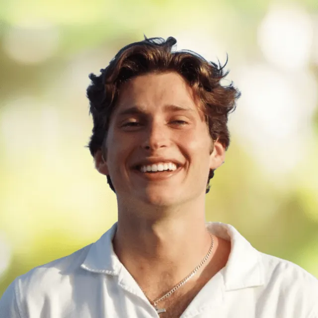 Smiling young man with wavy brown hair wearing a white shirt and silver chain against a blurred green background.