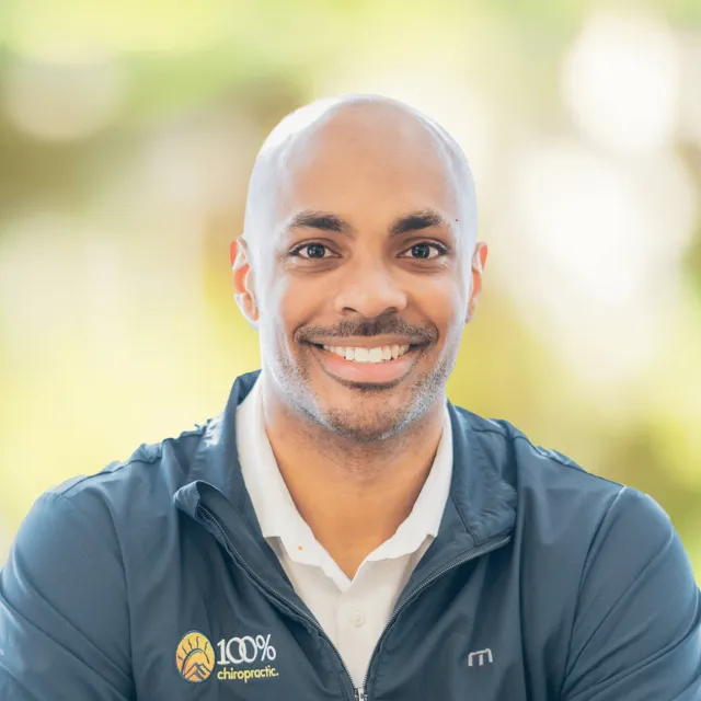 Smiling man wearing a 100% chiropractic jacket in an outdoor setting with soft natural light background