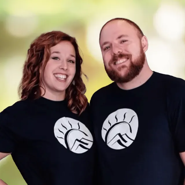 Smiling man and woman wearing matching black t-shirts with white mountain and sun logo standing side by side.