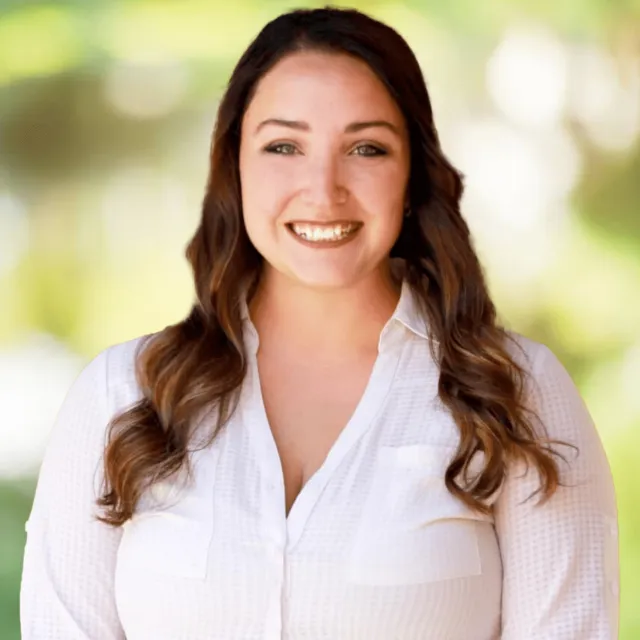 Smiling woman with long brown hair wearing a white shirt against a blurred green outdoor background