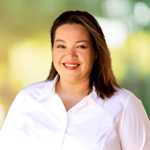 Smiling woman with medium-length dark hair wearing a white shirt against a blurred green background