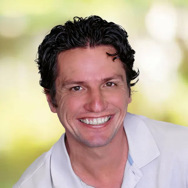 Smiling man with curly black hair in a white collared shirt against a soft green blurred background.