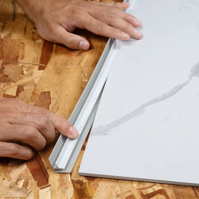 Hands installing white flexible trim strip on wood surface next to marble-patterned tile slab.