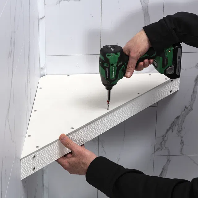 Installing a white triangular corner shelf with screws using a green and black cordless drill by a person.