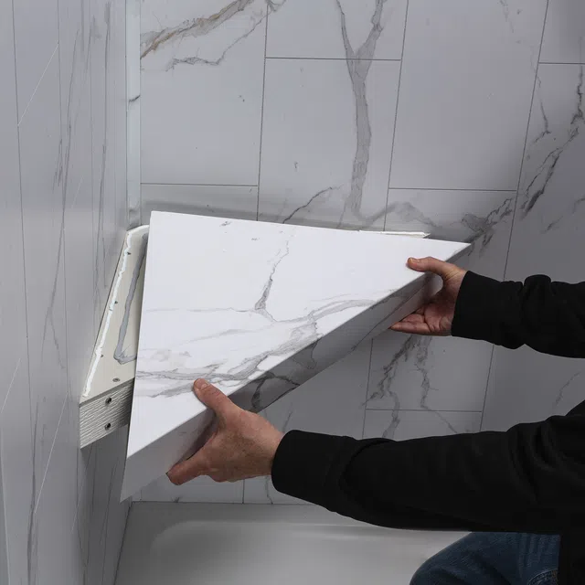 Hands installing a white marble-patterned wall panel over adhesive in a bathroom corner above a bathtub.