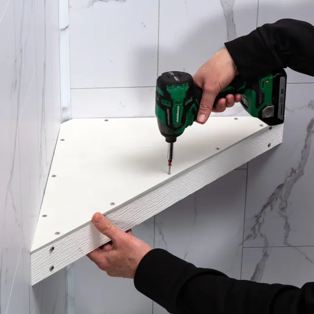 Person installing white wooden shelf with a green cordless drill on a marble tiled wall.