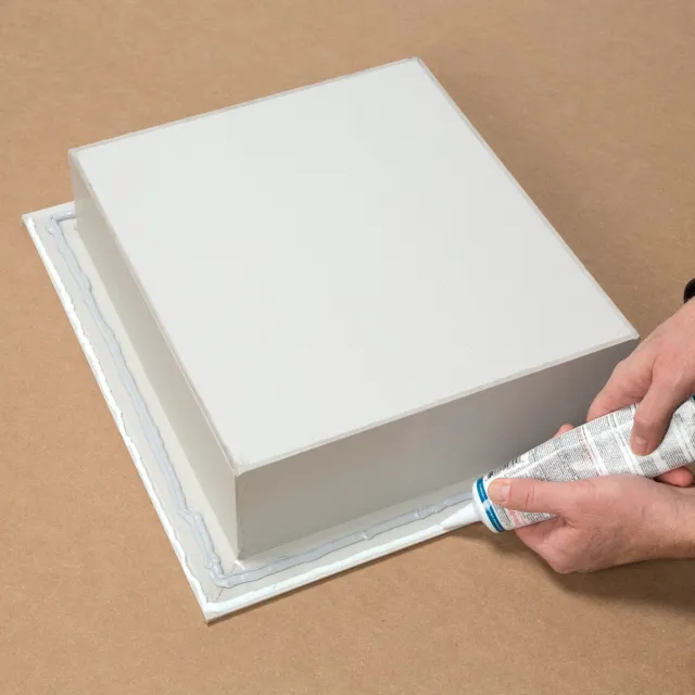 Hands applying adhesive from a tube around the base of a white cube on a brown surface for mounting.