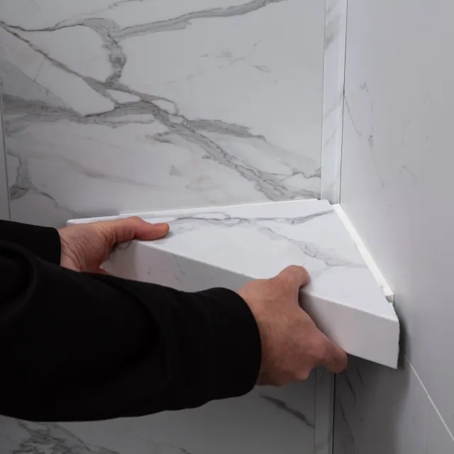Hands installing a white marble corner shelf against matching marble walls in a bathroom or kitchen setting.