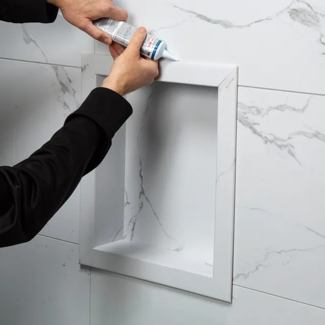 Person applying sealant to a white marble tiled bathroom niche for waterproofing and finishing.