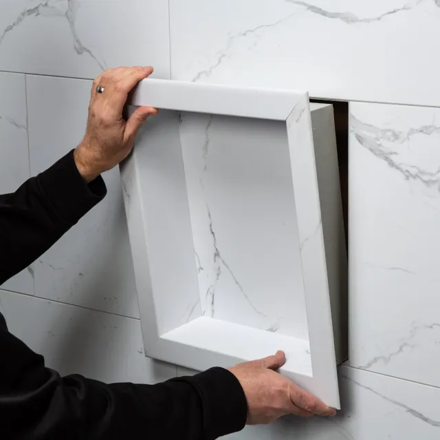 Hands installing a white marble recessed shower niche into a tiled wall with gray veining.