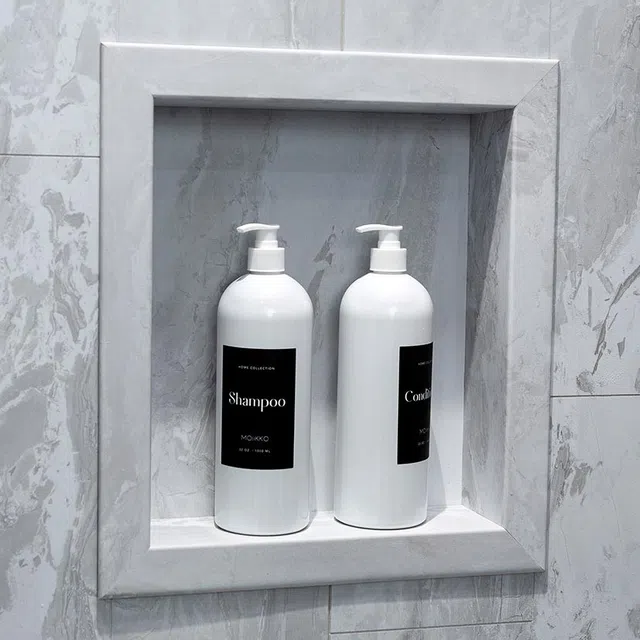 Two white pump bottles labeled shampoo and conditioner in a modern gray marble shower niche.