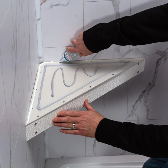 Person applying adhesive to a triangular shower shelf for bathroom tile installation with marble wall tiles.