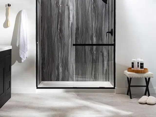 Modern bathroom with gray marble shower, black framed glass door, white towel, and bath accessories on stool.