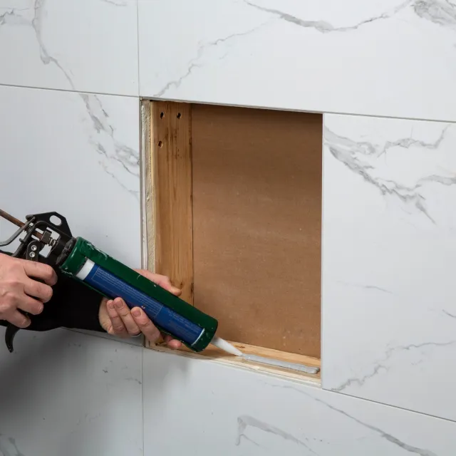 Person applying white sealant with caulking gun to a rectangular hole in marble-patterned wall tiles