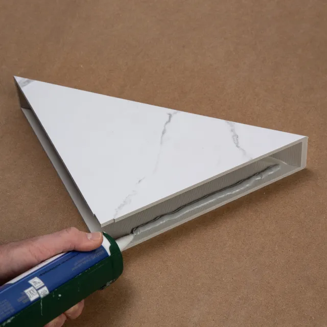 Hand applying white adhesive sealant to the edge of a triangular marble tile on a brown surface.