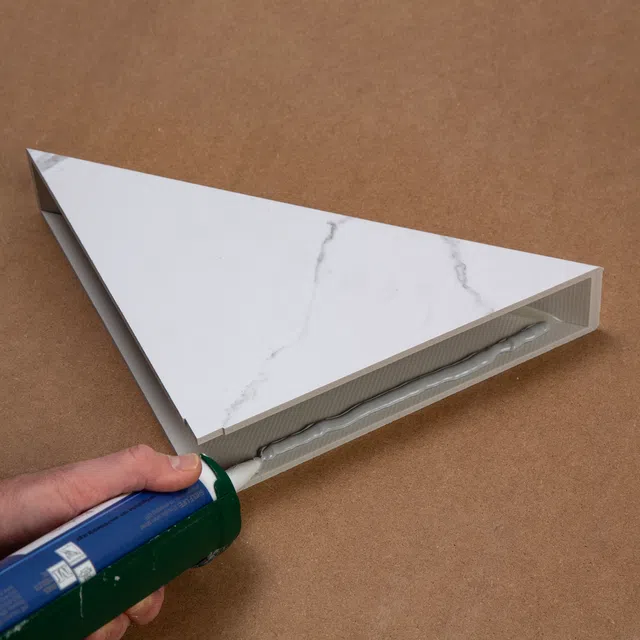 Applying adhesive sealant on the edge of a triangular marble-patterned tile on a brown surface.