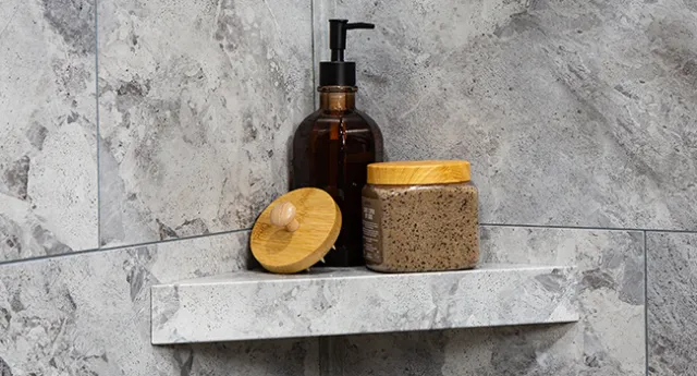 Amber pump bottle and natural scrub jar with wooden lid on gray stone shower shelf corner.