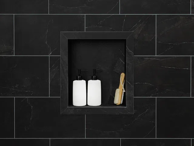 Two white pump bottles and a wooden brush in a black tiled wall niche in a modern bathroom.