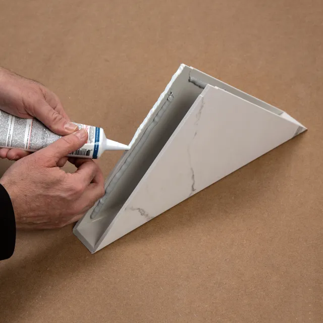 Person applying adhesive to a white marble-patterned triangular shelf on a brown surface.