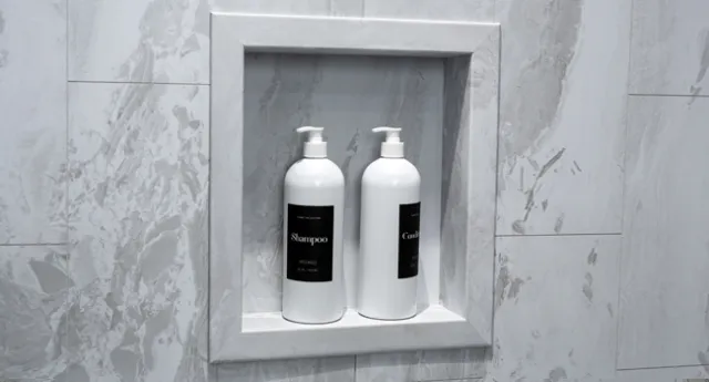 Two white shampoo and conditioner bottles with black labels on a gray marble shower niche shelf.