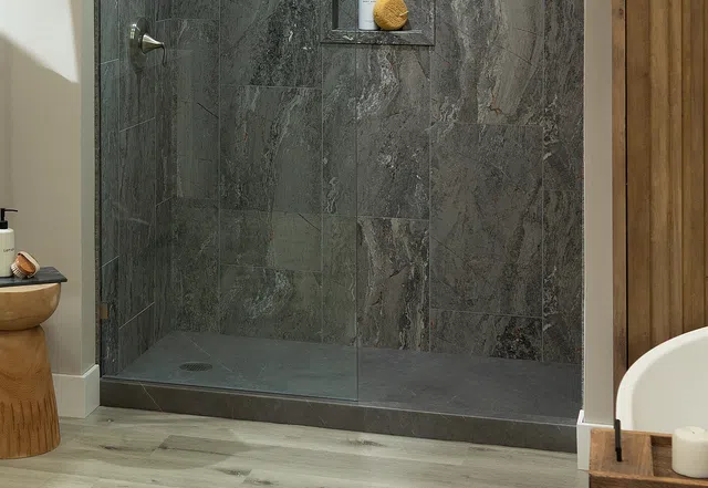 Modern bathroom shower with dark gray stone tiles, glass door, and light wood flooring and accents
