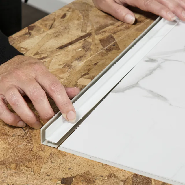 Hands installing a white tile edge trim on a marble-patterned floor tile over plywood subfloor.