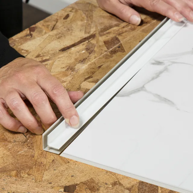 Hands installing white tile trim on wood surface next to white marble tile with grey veining.