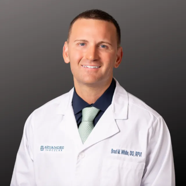 Smiling male doctor in white coat with Advanced Vascular logo and green tie on gray background