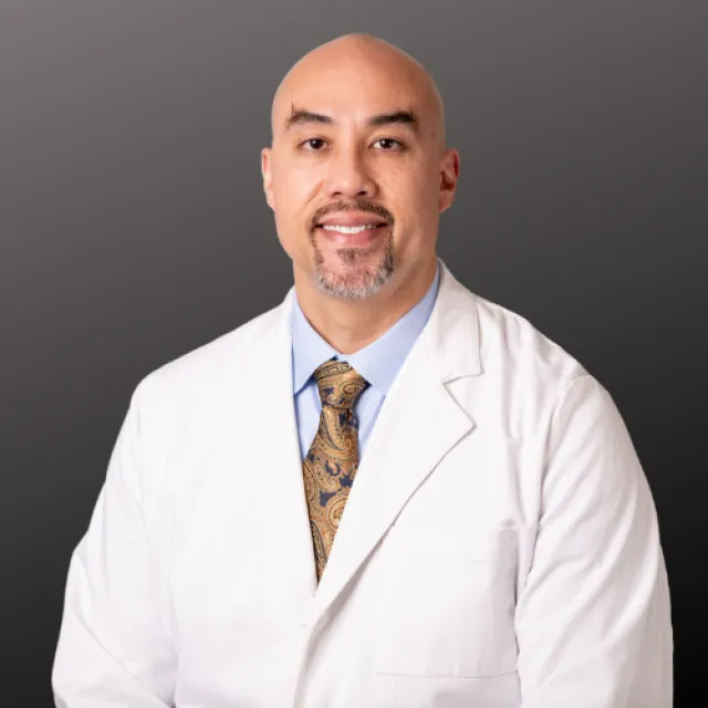 Smiling male doctor in white coat with blue shirt and patterned tie against gray background