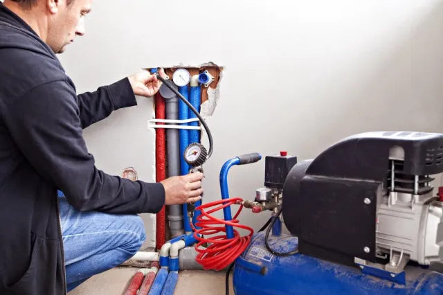Technician checking water pressure gauges on pipes connected to a blue air compressor in a wall cavity.