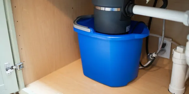 Blue bucket placed under kitchen sink pipes and garbage disposal unit inside wooden cabinet.