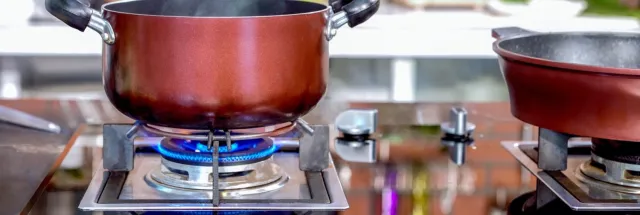 Red pot steaming on a gas stove burner with blue flame, next to a second pot on a reflective kitchen countertop.