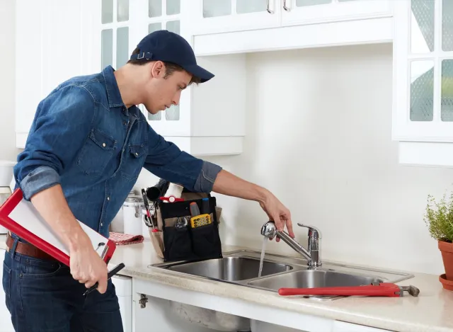 Plumber inspecting kitchen sink faucet with toolbox and wrench on countertop in modern white kitchen