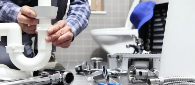 Plumber assembling white PVC pipe under a sink amid plumbing tools and parts in a tiled bathroom.