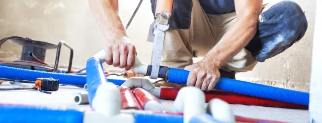 Plumber working with blue and red pipes using a cutting tool on a concrete floor in construction site.