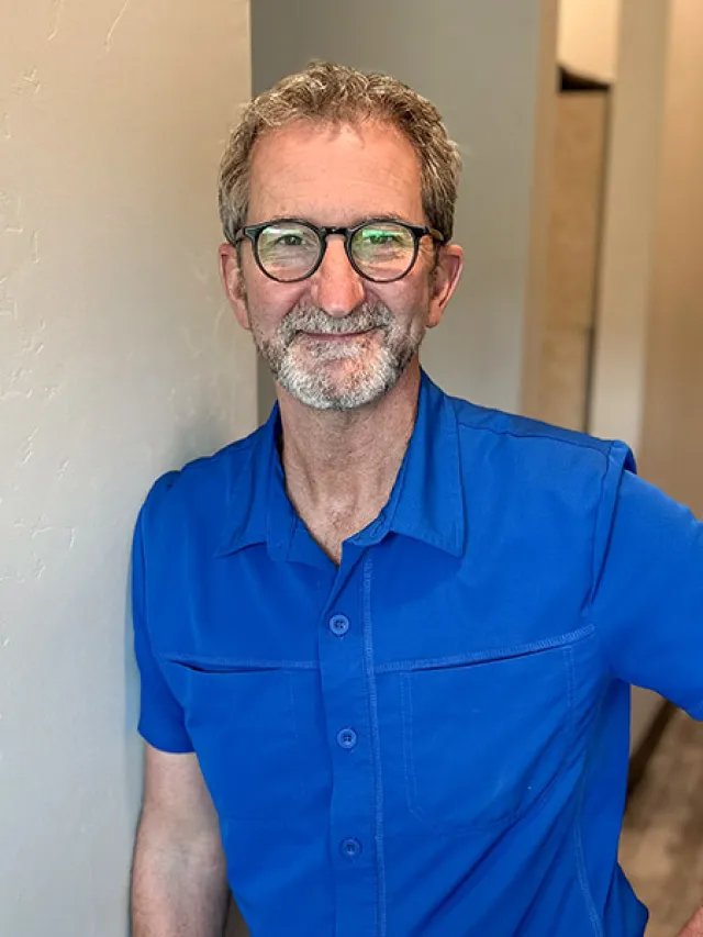Smiling middle-aged man with glasses and beard wearing a bright blue shirt leaning against a wall indoors.