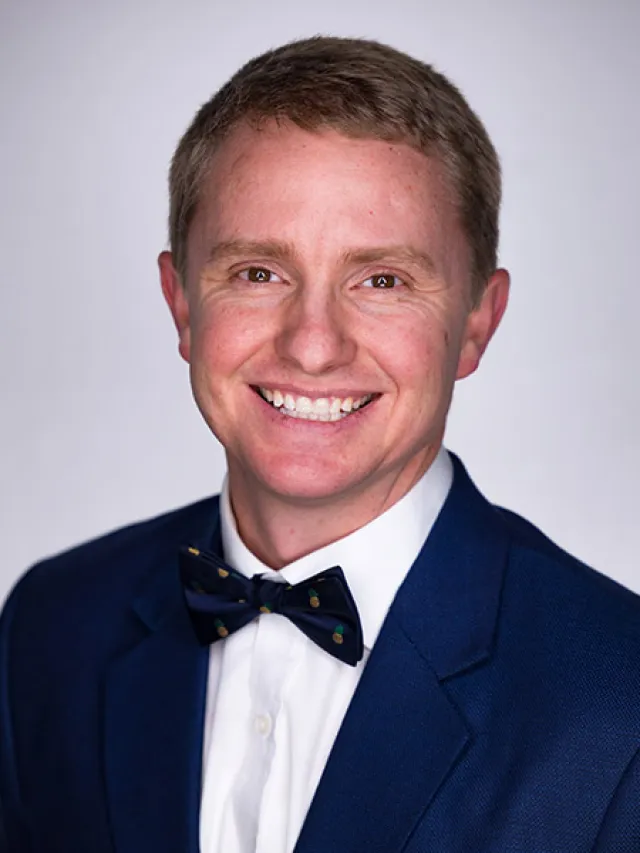 Smiling man in blue suit jacket, white shirt, and patterned bow tie against a neutral gray background.