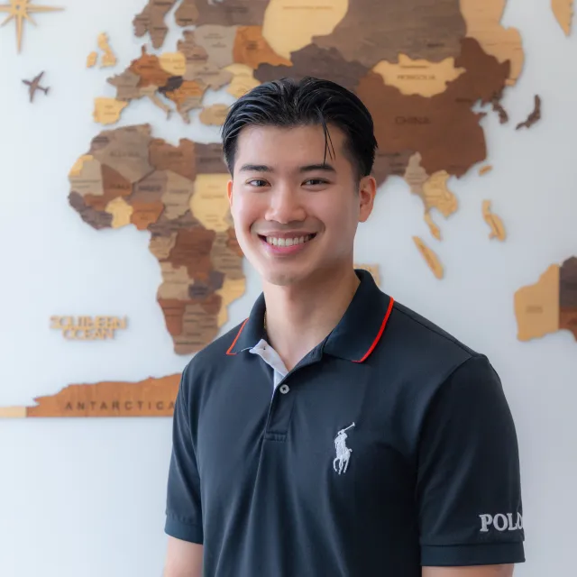 Smiling young man in black Polo shirt standing in front of a wooden world map on white wall.