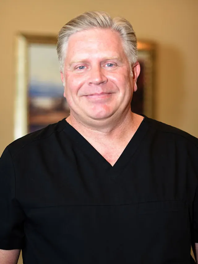 Smiling male medical professional in black scrubs against a neutral background.