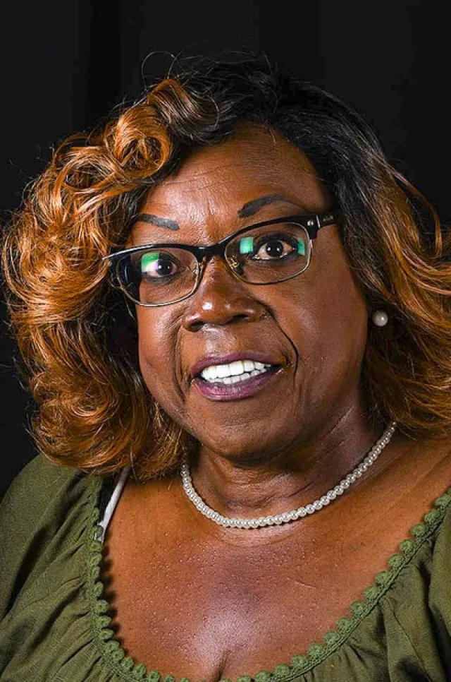 Smiling middle-aged black woman with glasses, curly hair, green top, pearl necklace, and earrings on black background.