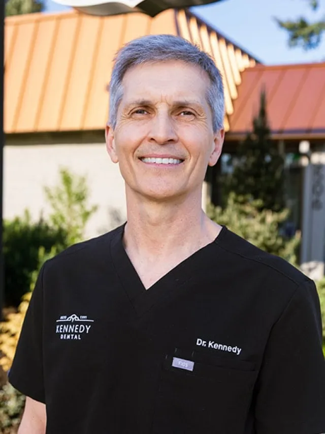 Smiling male dentist Dr. Kennedy in black scrubs standing outdoors near a building with an orange roof.