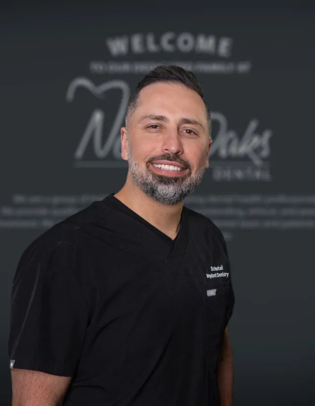 Smiling male dentist in black scrubs standing in front of a blurred dental clinic welcome sign.