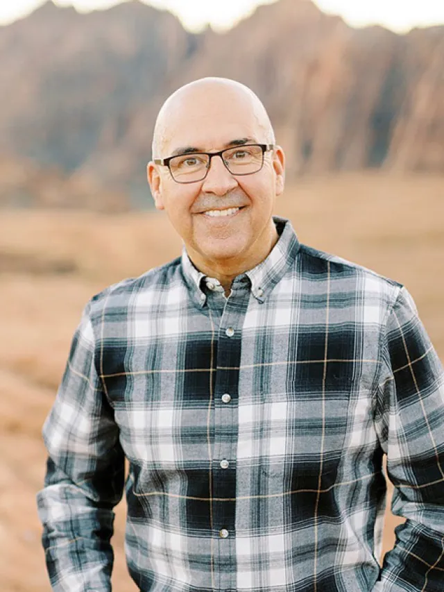 Smiling bald man wearing glasses and a plaid shirt standing outdoors with mountains in the background
