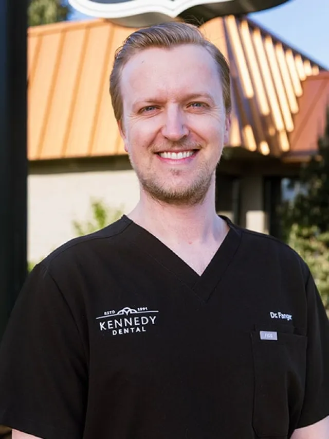 Smiling male dental professional wearing black Kennedy Dental scrubs outdoors in front of building.