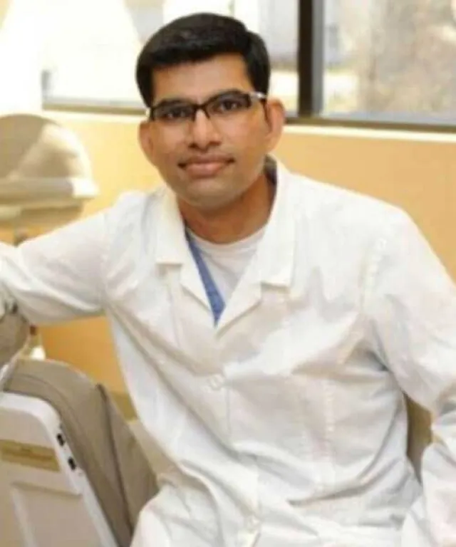 A dentist in a white coat smiles while seated in a dental office setting.