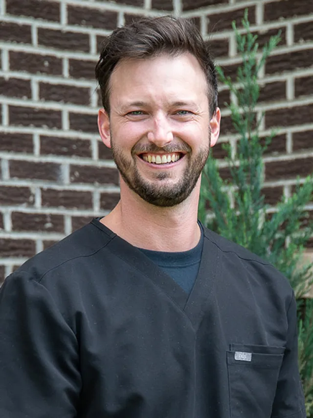 Smiling man with short brown hair and beard wearing black scrub top against brick wall and greenery background