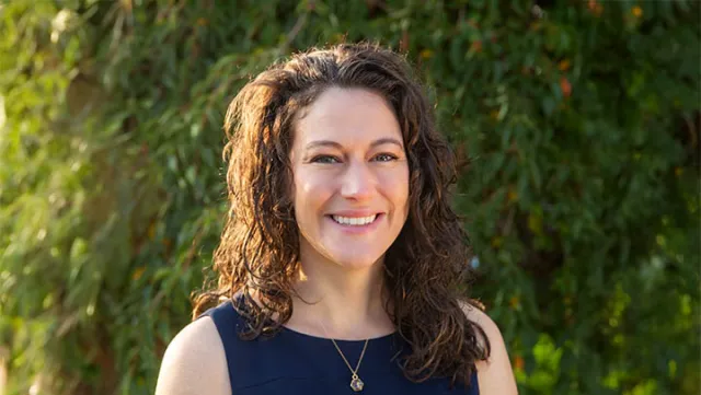 Smiling woman with curly brown hair wearing a navy blue sleeveless top standing outdoors with greenery background
