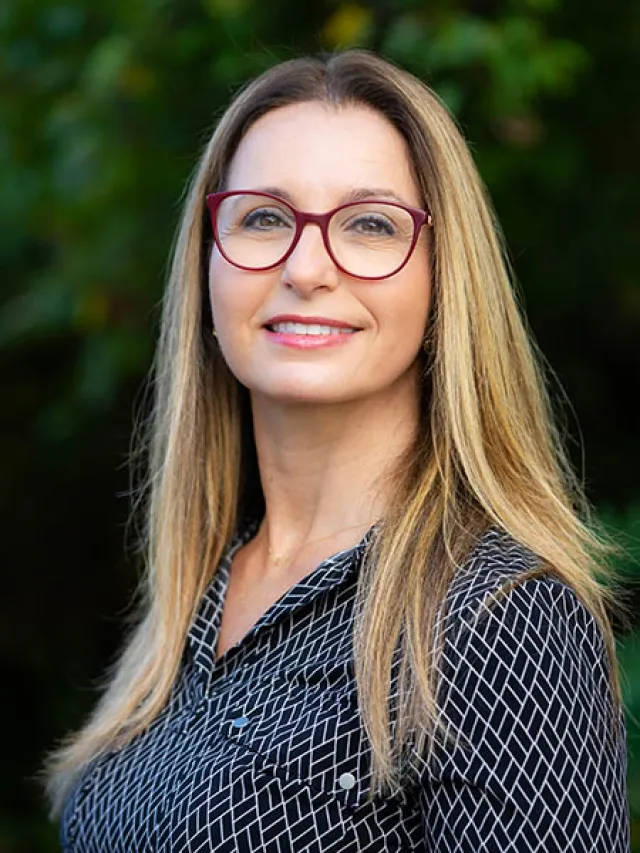 Smiling woman with long blonde hair wearing red glasses and a black patterned shirt outdoors.